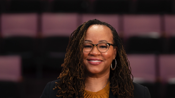 A headshot of Rev. Dr. Shonda Jones, a Black woman with shoulder-length hair, black glasses, and silver earrings. She is smiling at the camera in front of a black and purple background. 