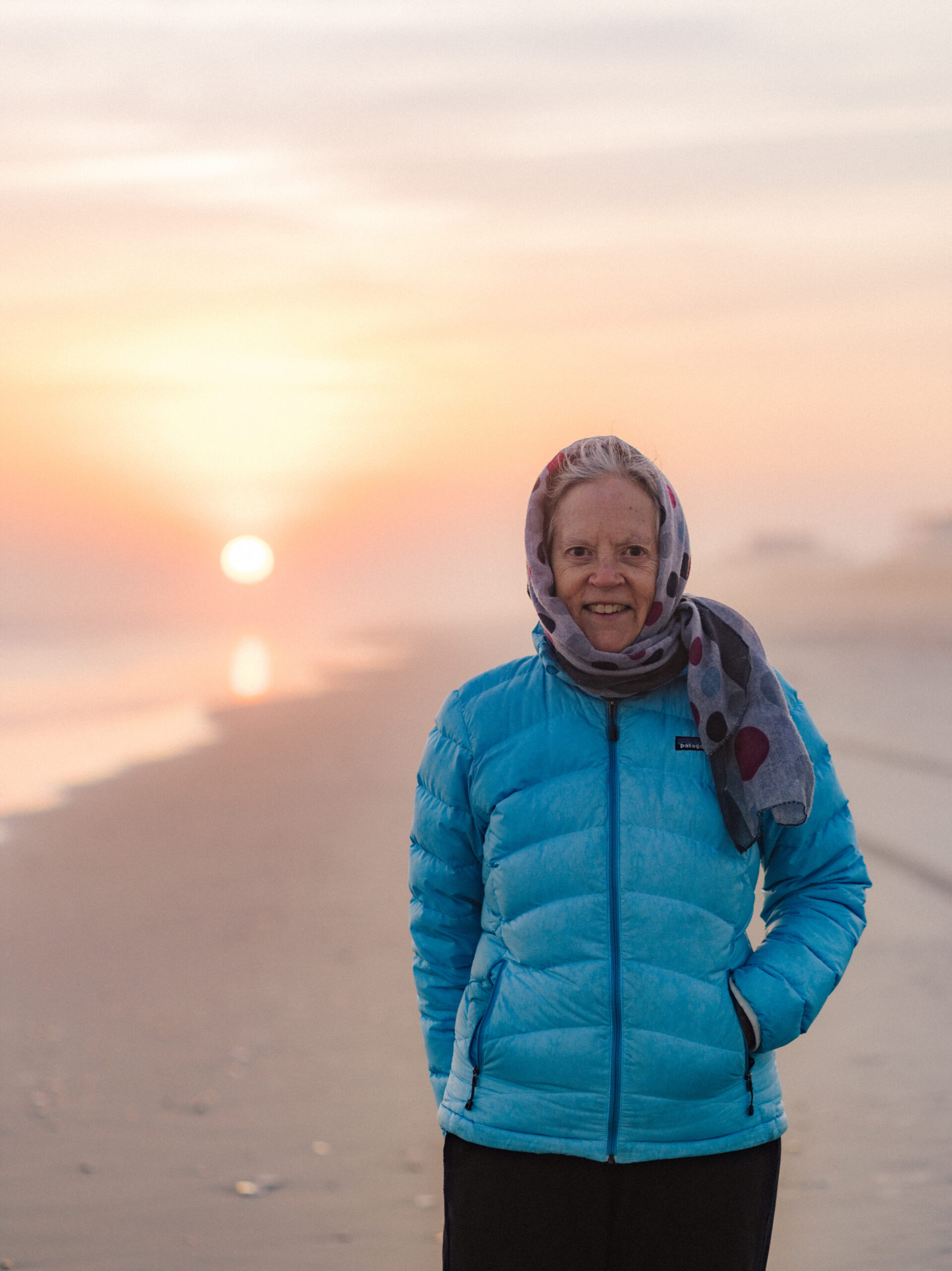 Jeanette Stokes smiles at the camera while standing on the beach with a jacket and scarf on